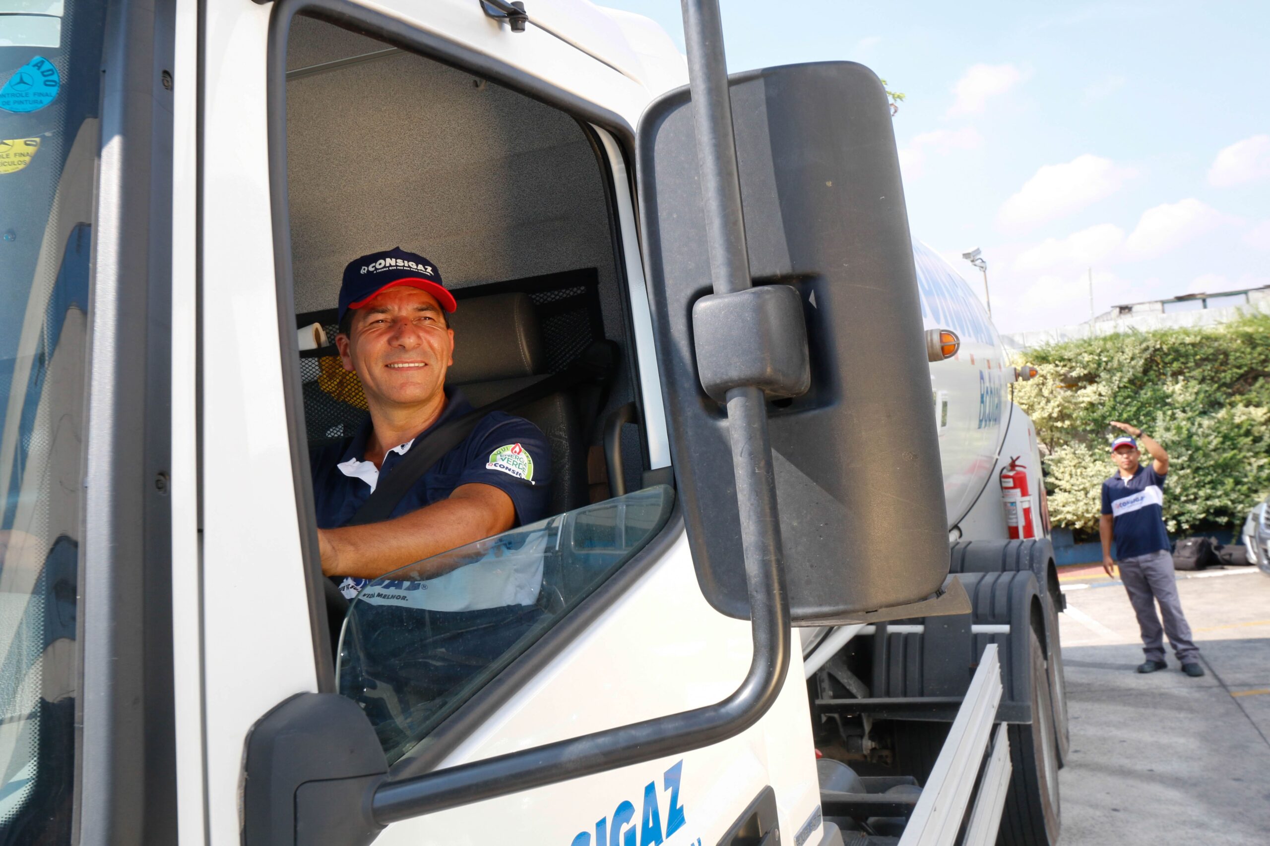 Motorista de caminhão de gás sorrindo e acenando, com outro trabalhador ao fundo, durante o dia em área de estacionamento ao ar livre. Para saber como contratar funcionário