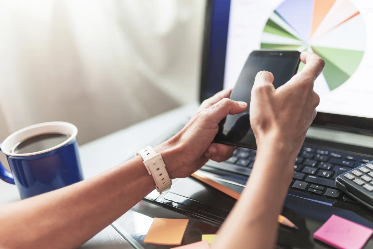 Pessoa usando smartphone em frente a laptop com gráfico de pizza na tela, xícara de café e calculadora na mesa, aprendendo como divulgar minha empresa gratuitamente