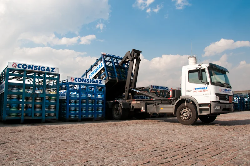 Caminhão branco transportando botijões de gás em estrutura metálica azul da Consigaz em pátio com céu azul e nuvens, retratando a logística de entrega dos botijões.
