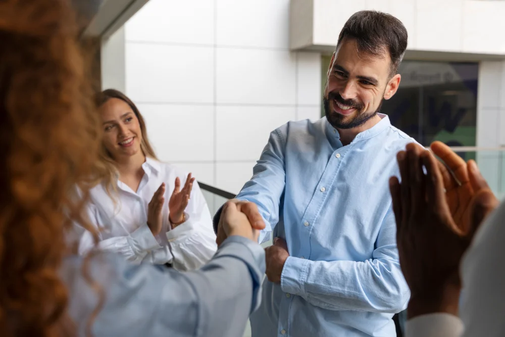 Homem sorridente apertando a mão de outra pessoa em ambiente de escritório com colegas aplaudindo ao fundo, demonstrando o sucesso das parcerias certas.
