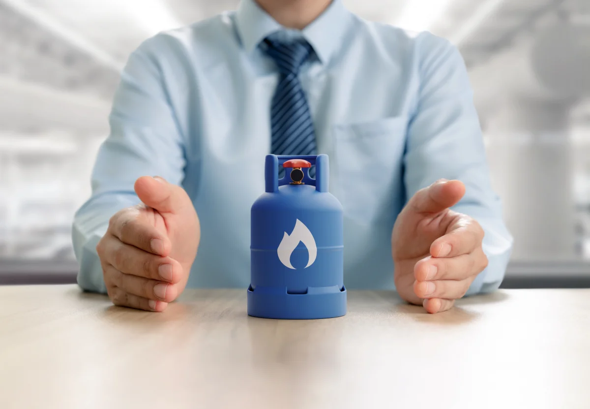 Botijão de gás azul com símbolo de chama protegido por mãos de homem de camisa azul, representando o seguro empresarial.