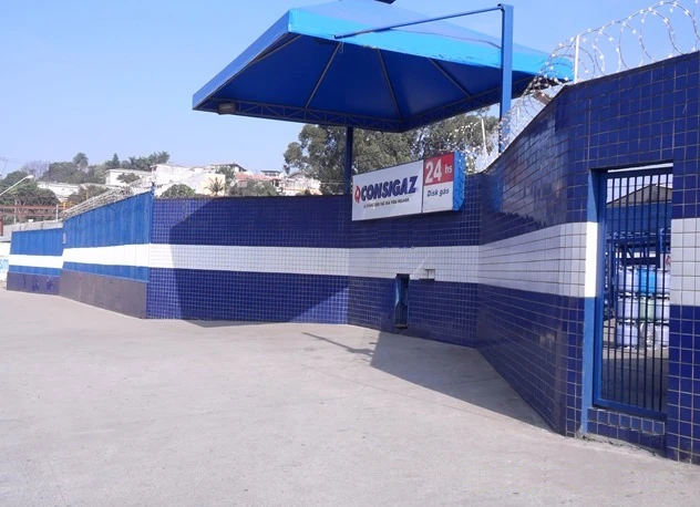 Fachada de uma loja de gás em cores azuis e brancas com um toldo azul e uma parede de azulejos, com placa de sinalização de venda de gás de 24 horas.