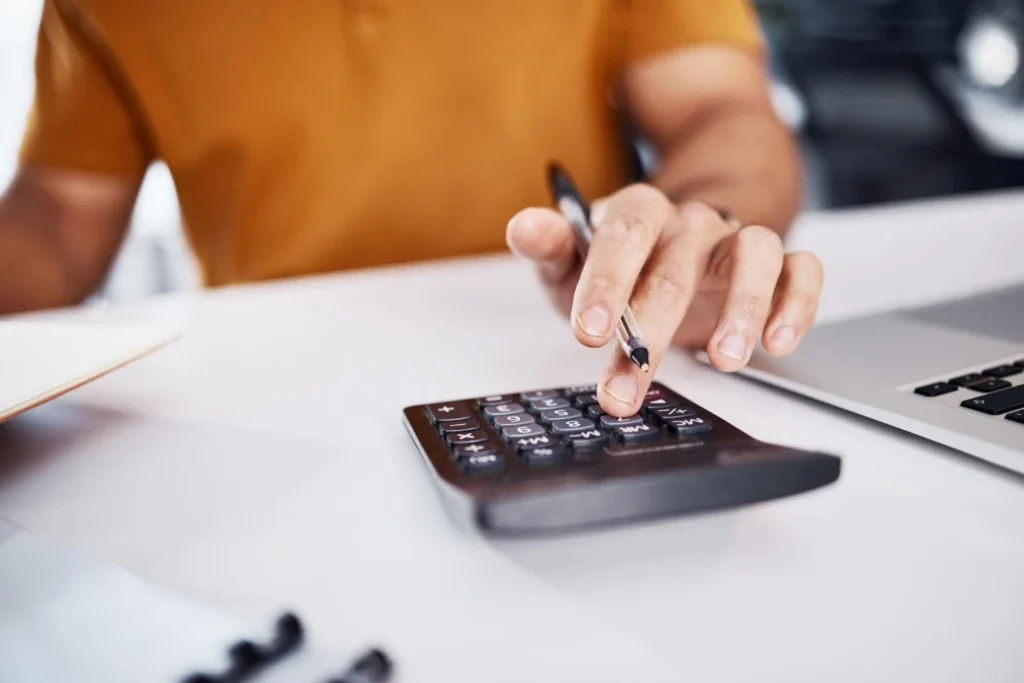Pessoa em mesa de trabalho utilizando calculadora e caneta para registrar dados financeiros, em ambiente de escritório, com foco na contagem de valores e controle de custos, franquia sem royalties
