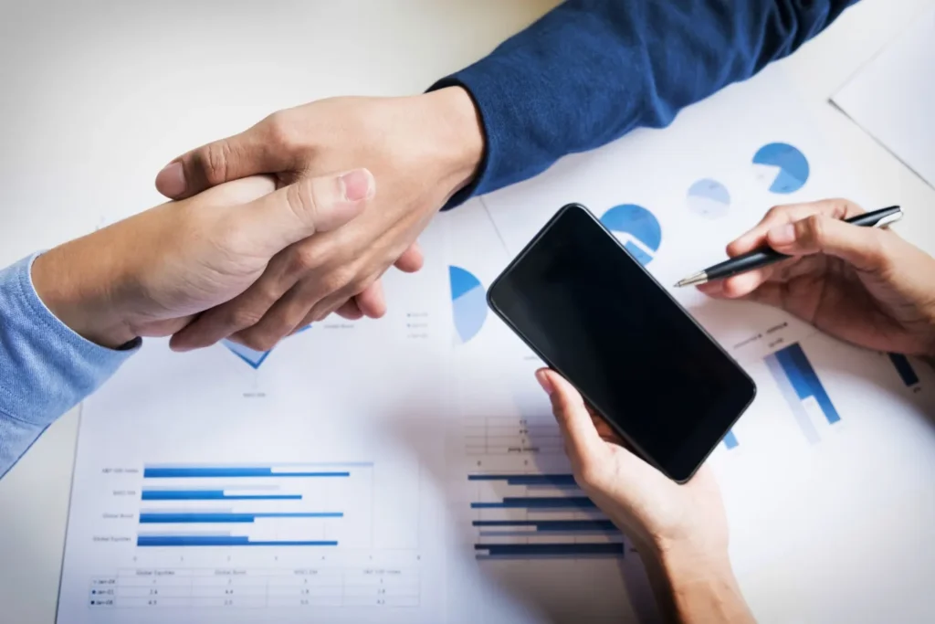 Mãos em uma reunião analisando gráficos e indicadores de desempenho em relatórios, com smartphone e caneta sobre a mesa, sugerindo consultoria de negócio
