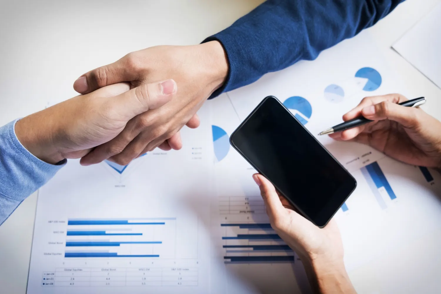 Mãos em uma reunião analisando gráficos e indicadores de desempenho em relatórios, com smartphone e caneta sobre a mesa, sugerindo consultoria de negócio